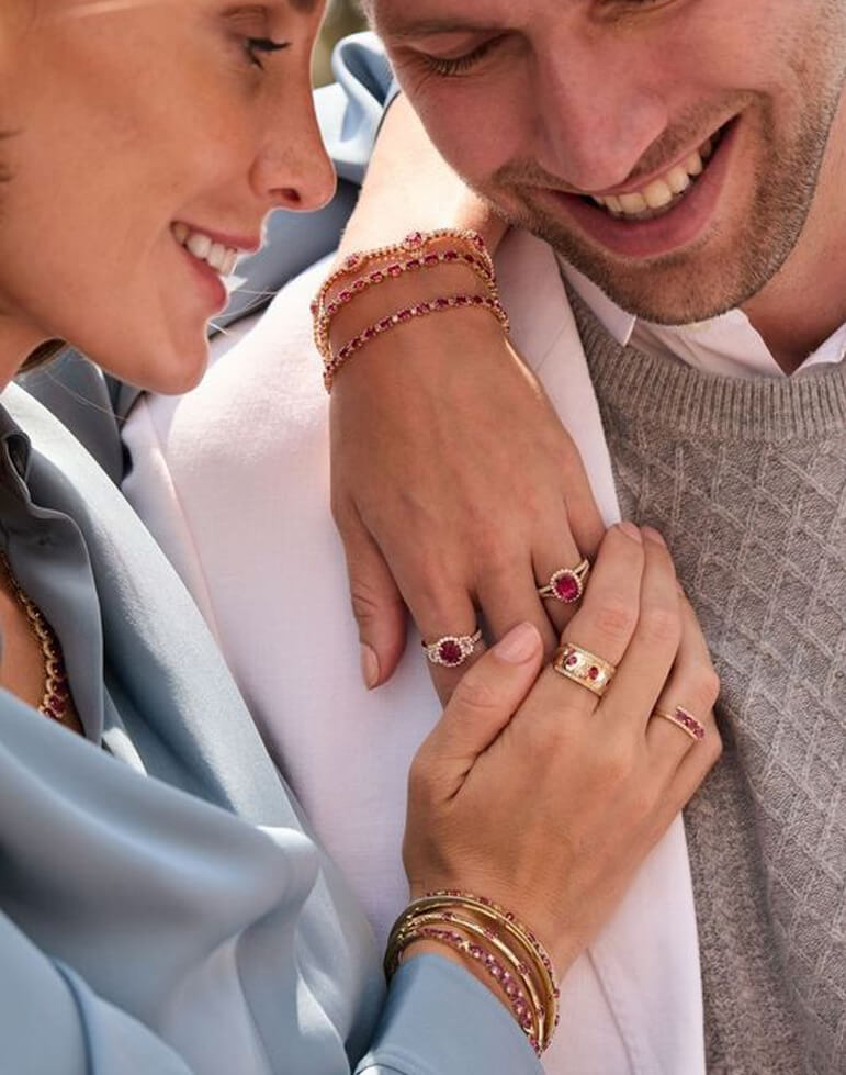 Smiling couple displaying a romantic array of Le Vian jewelry, including ruby and pink sapphire rings, bracelets, and bangles in yellow and rose gold, symbolizing love and connection.