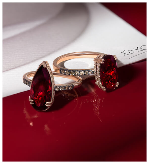 Two Le Vian rings with deep red garents in pear and oval cuts, set in rose gold with Chocolate Diamond® pavé bands, placed on a red lacquered surface and white envelope.