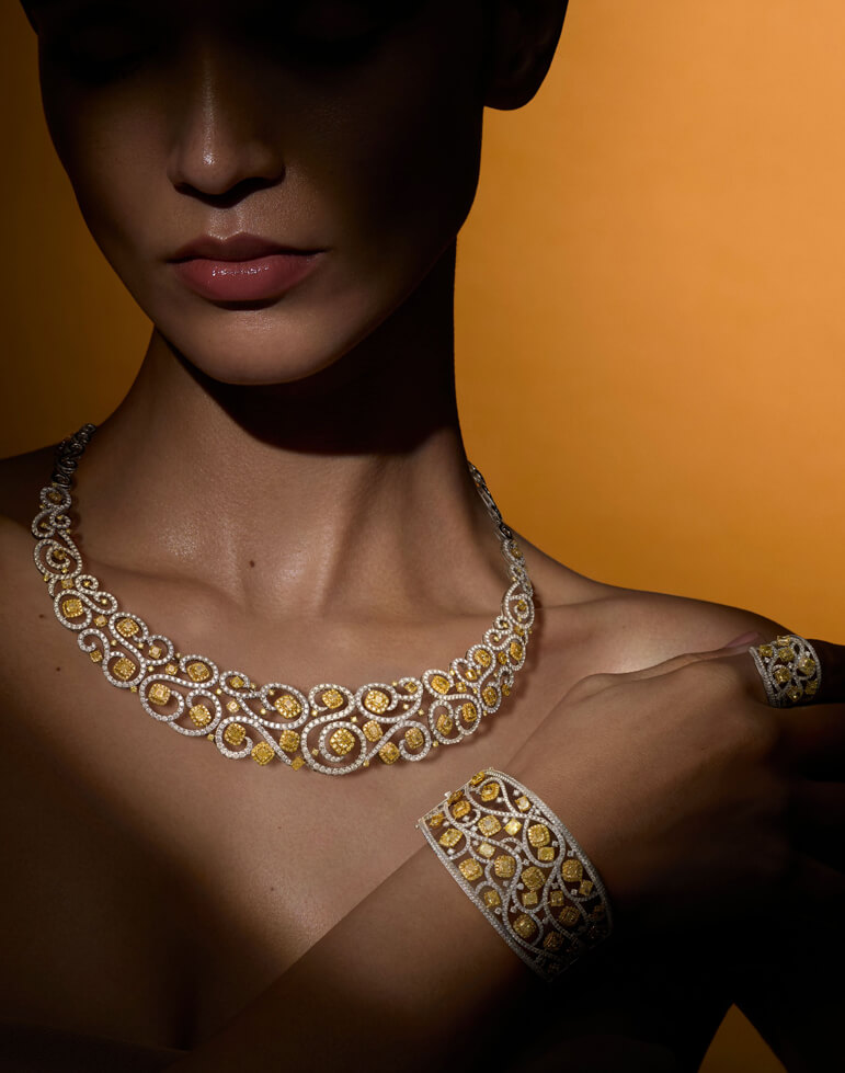 Elegant woman wearing a high jewelry suite from Le Vian featuring a necklace, bracelet, and ring with yellow diamonds and intricate white diamond filigree in an ornate, lace-like design.