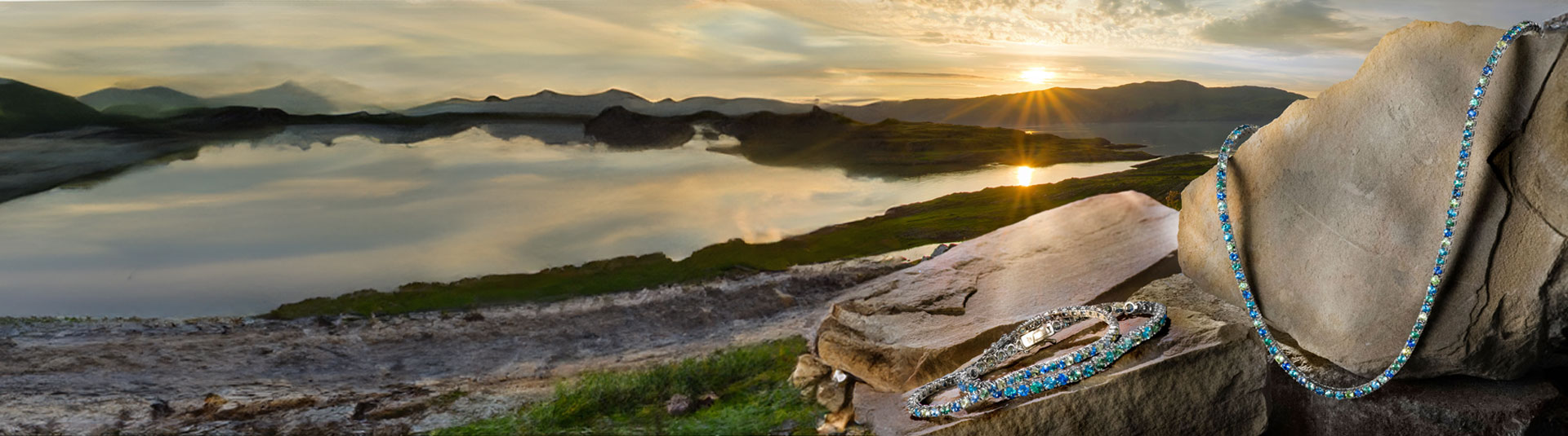 Le Vian's 2026 jewelry collection featuring ombré blue and green gemstone bracelet and necklace, styled on natural stone with a scenic mountain lake at sunset in the background.
