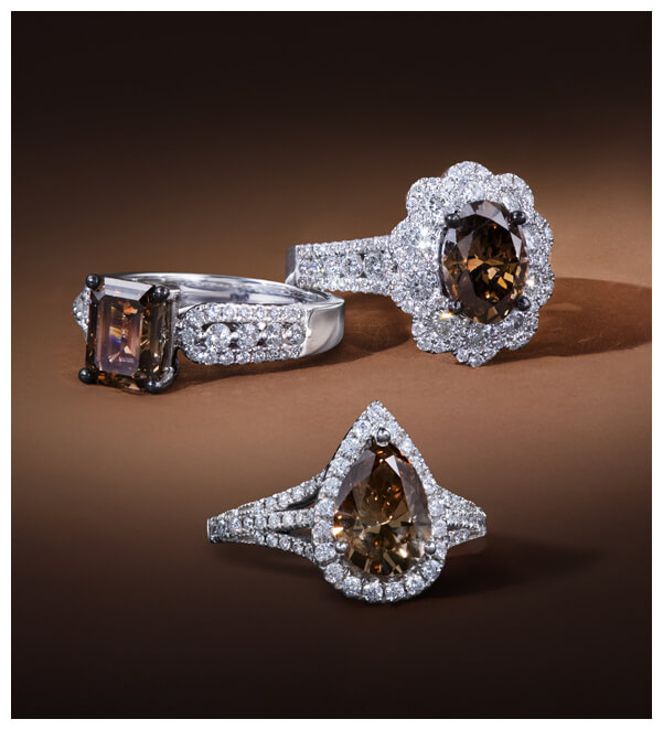 Trio of Le Vian Chocolate Diamond® rings in white gold settings, showcasing emerald, oval, and pear-shaped brown diamonds surrounded by pavé white diamonds on a chocolate-hued background.