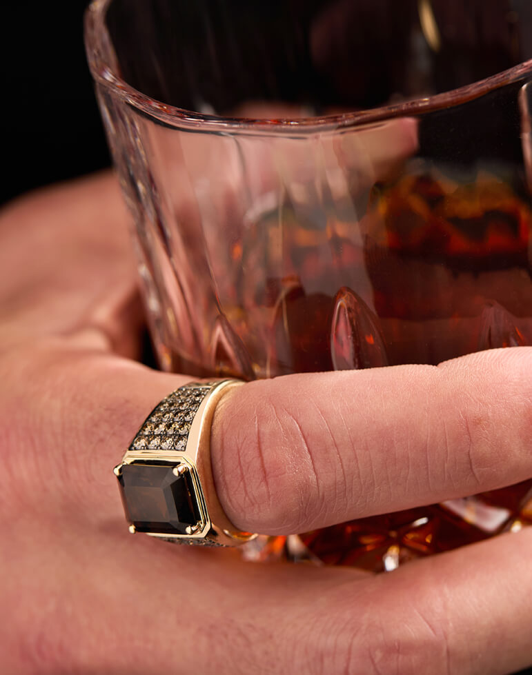 Close-up of a man's hand holding a whiskey glass while wearing a bold Le Vian men’s ring featuring a rectangular Chocolate Diamond® in a yellow gold setting with pavé accents.