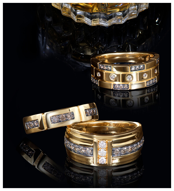 Trio of Le Vian men’s rings in polished yellow gold with bold architectural designs and pavé set Chocolate Diamonds® and white diamonds, displayed on a glossy black surface.