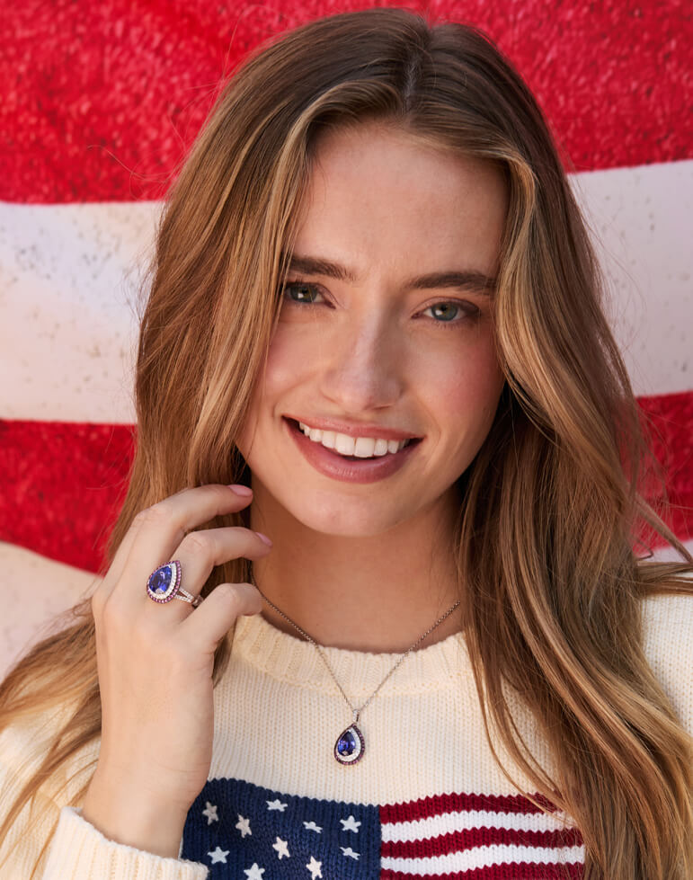 Woman wearing patriotic fine jewelry with a pear-shaped blue gemstone ring and matching pendant necklace, posing in front of an American flag.