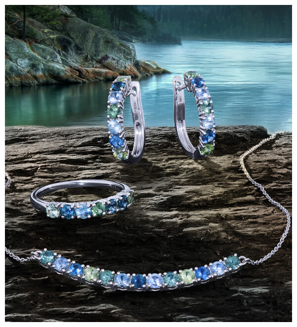 Close-up of Le Vian Americana collection jewelry, featuring a Montana Sapphire blue stone ring, hoop earrings, and necklace on a slate rock with a sunset.