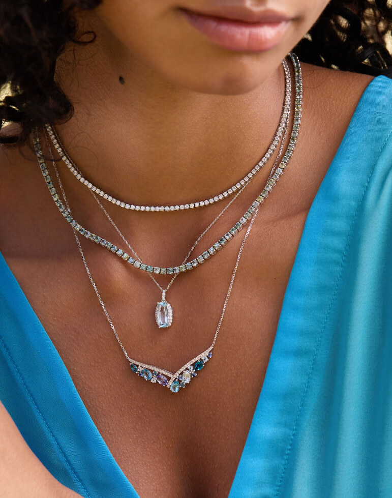 Woman wearing layered Le Vian gemstone necklaces, featuring aquamarine, Austrailian opal, pastel ombré gems, and a pear-shaped pendant, styled with a vibrant blue dress for an ocean-inspired look.