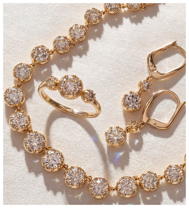 Matching yellow gold diamond bracelet, ring and earrings with round white diamonds in bezel-style settings, arranged on white textured fabric.