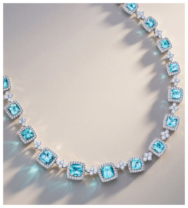 White gold necklace with cushion-cut turquoise-colored gemstones and white diamond accents arranged in a curved design on a neutral background.